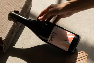Una mano tiene ferma una bottiglia di vino con l'etichetta 'GRIECI' su una superficie di cemento illuminata dal sole.